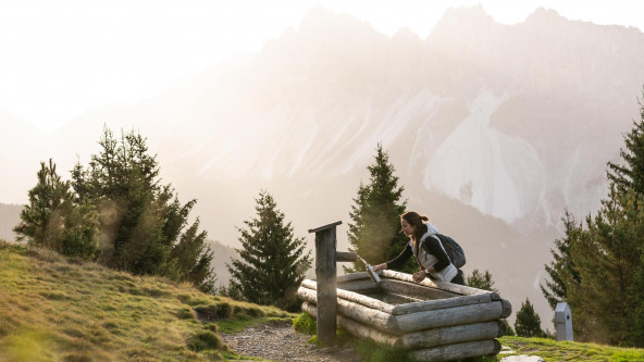 Una donna riempie la sua borraccia con acqua fresca da una fontana di legno situata in montagna, circondata da pini e con una vista mozzafiato sulle montagne al tramonto.