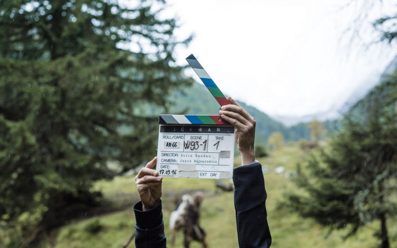 A film clapperboard can be seen in the middle of the picture. Behind it is a movie scene in a rainy forest.